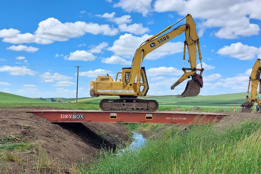 An excavator parked on a flat rack container bridge over an irrigation ditch