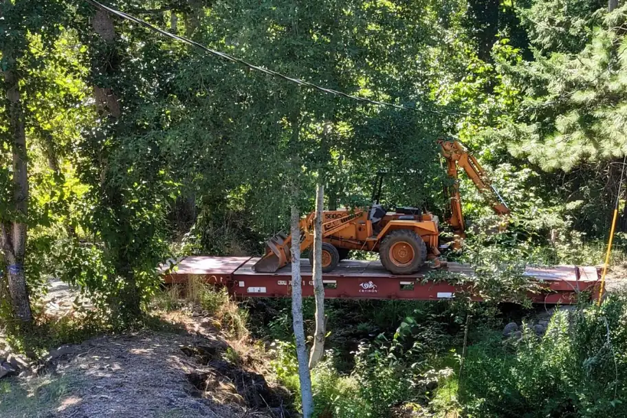 An orange tractor on a flat rack container bridge over a creek in the woods