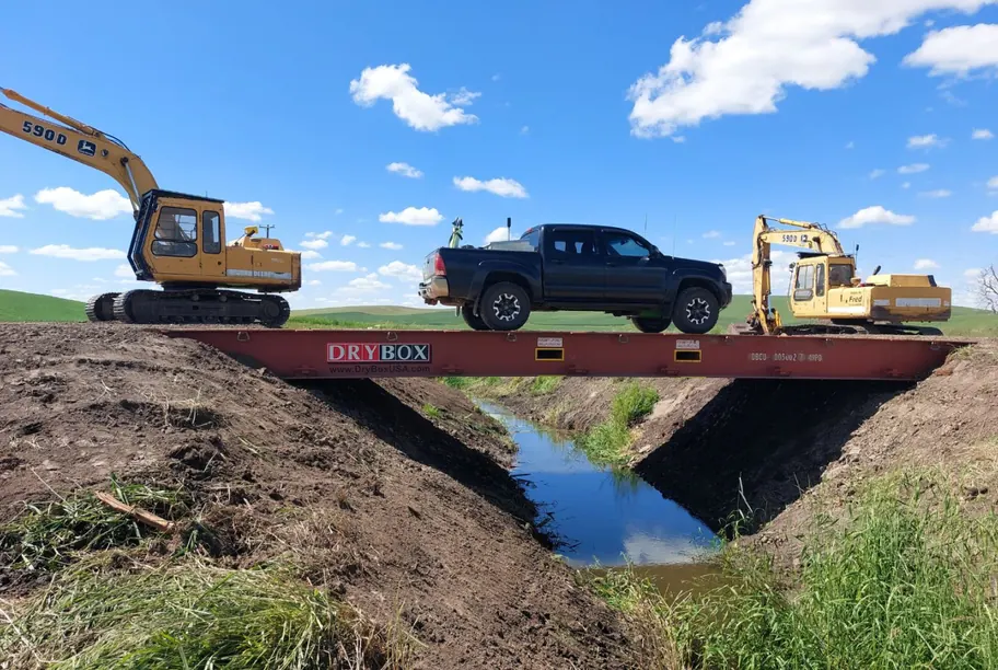 A pickup truck crossing a flat rack container bridge over an irrigation ditch