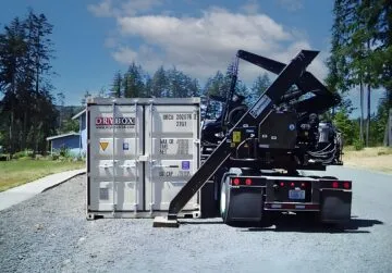 A beige shipping container on the ground next to side loader