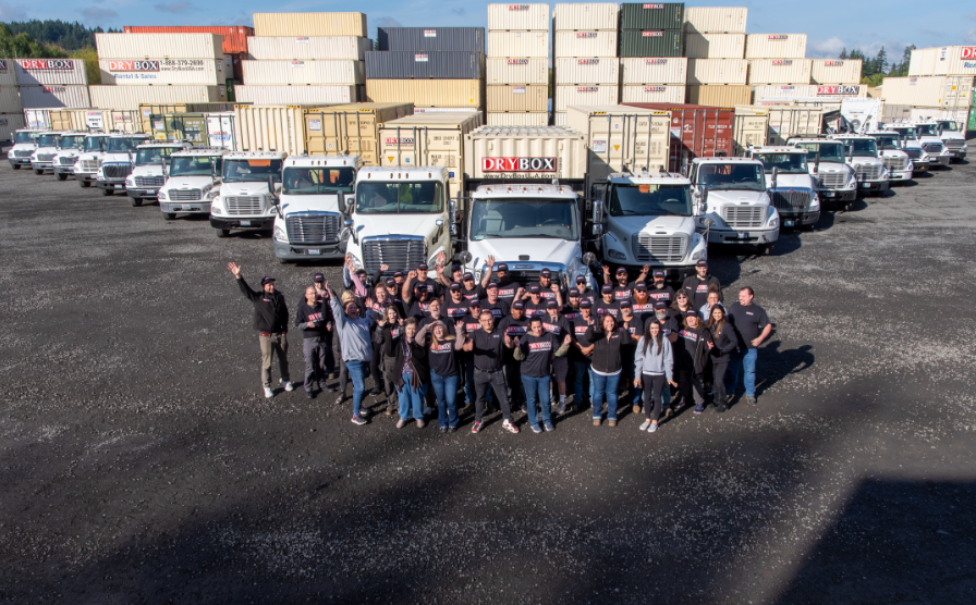 Employees standing in front of Dry Box trucks with a wall of containers in the background.